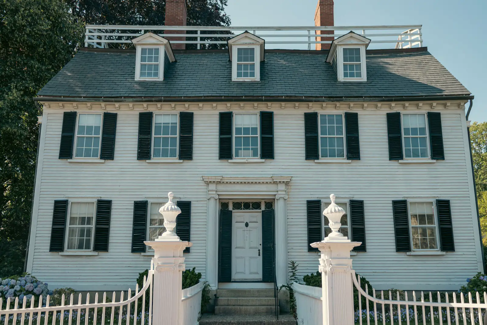 A white Georgian colonial house with black shutters, a balustraded roof and a white picket fence